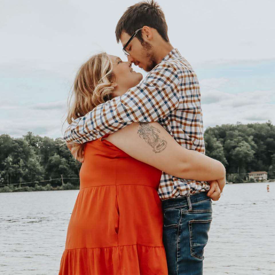 Couple embracing lakeside, woman in orange dress and man in plaid shirt, about to kiss with water and trees behind them