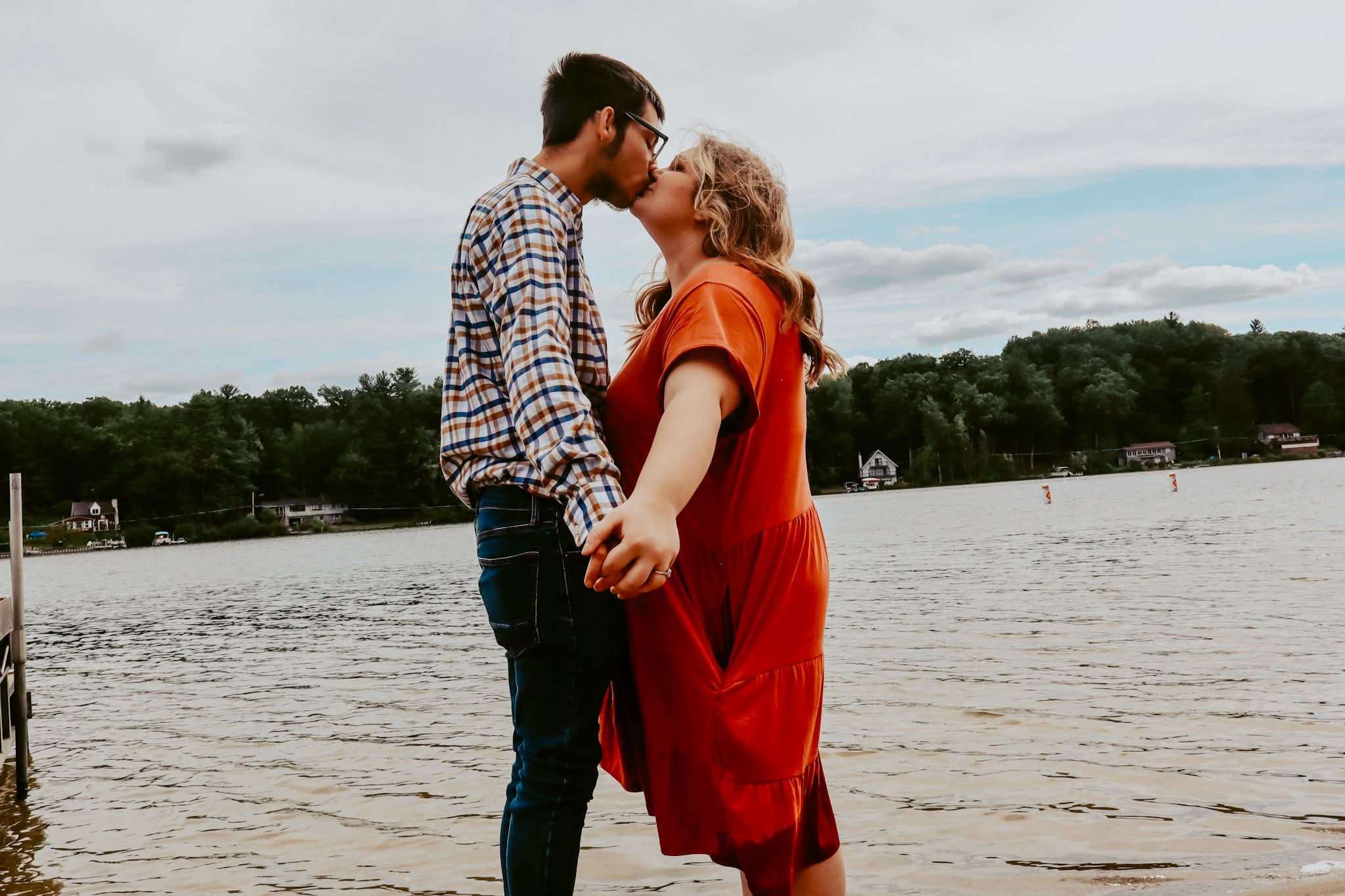 Couple in orange dress and plaid shirt kissing on the lakefront, overcast sky and treeline behind them