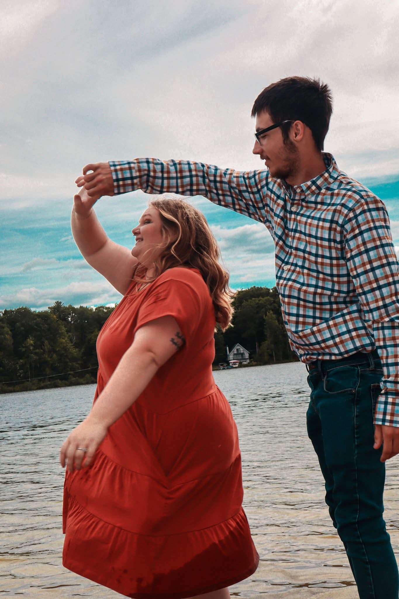 Man in plaid shirt spinning his partner in an orange dress by the lake, both laughing, dramatic cloudy sky above