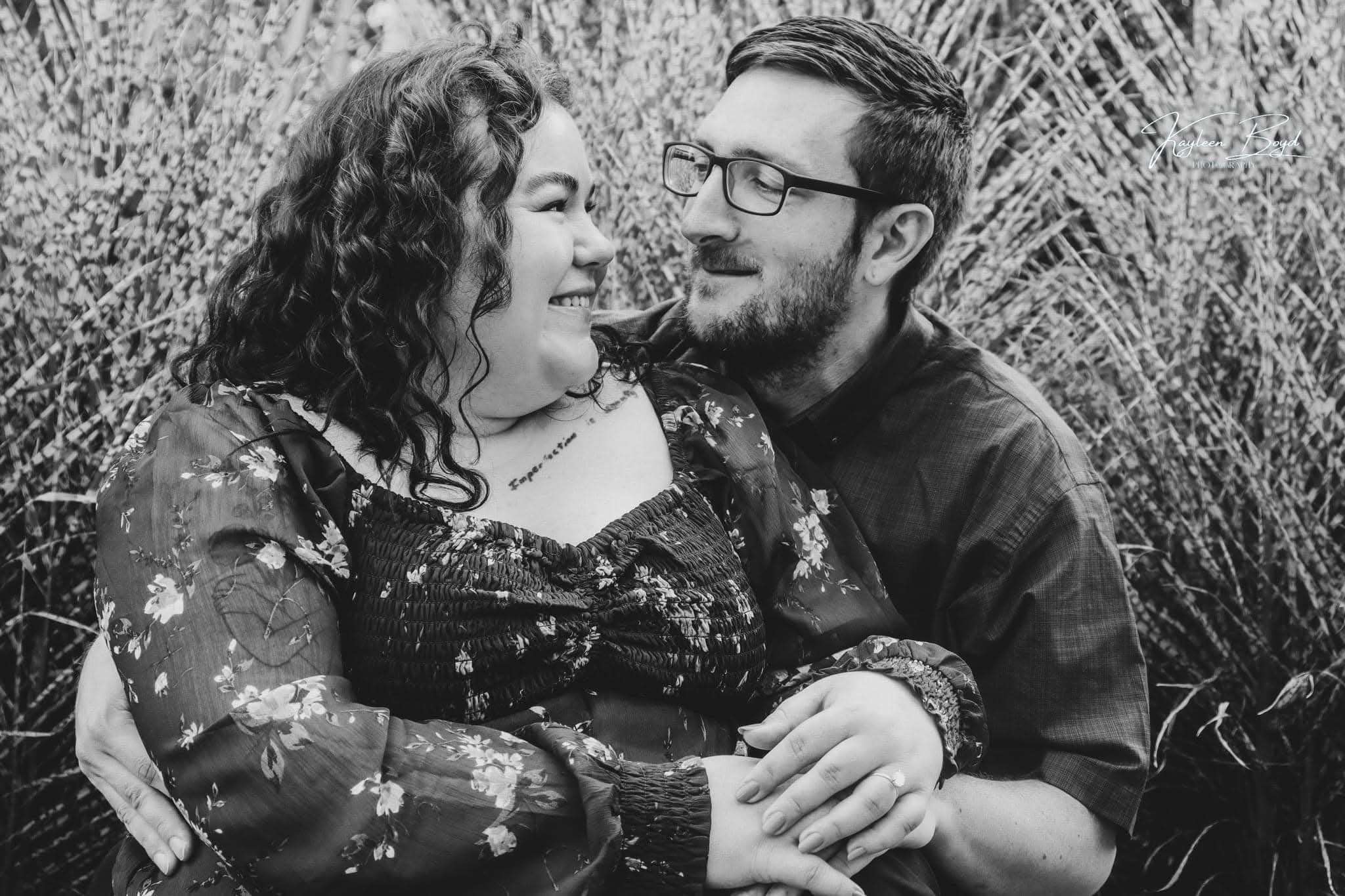 Black and white close portrait of engaged couple, woman leaning into her partner, engagement ring on her hand