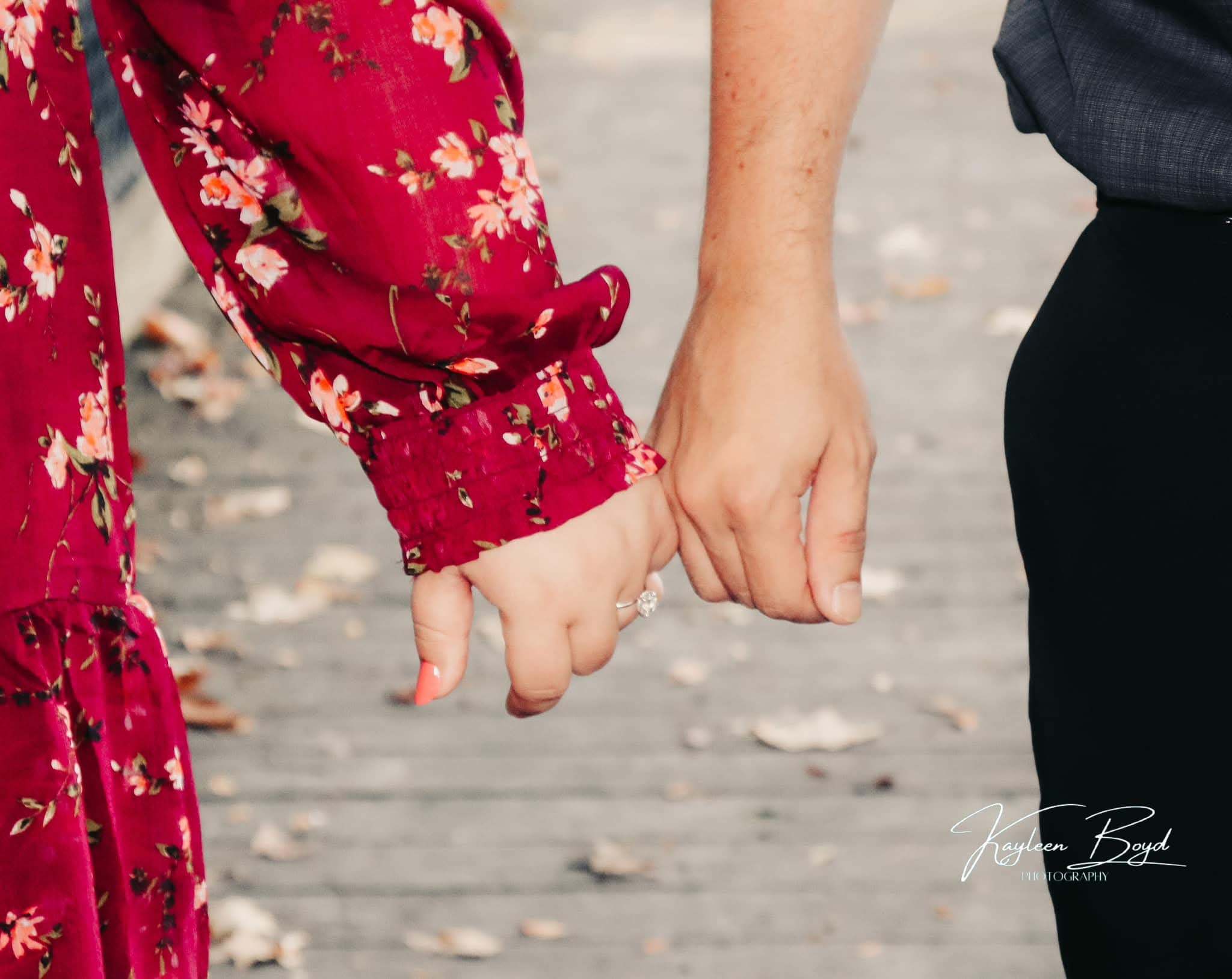 Close-up of couple holding hands on a boardwalk, engagement ring prominent, fallen leaves beneath them