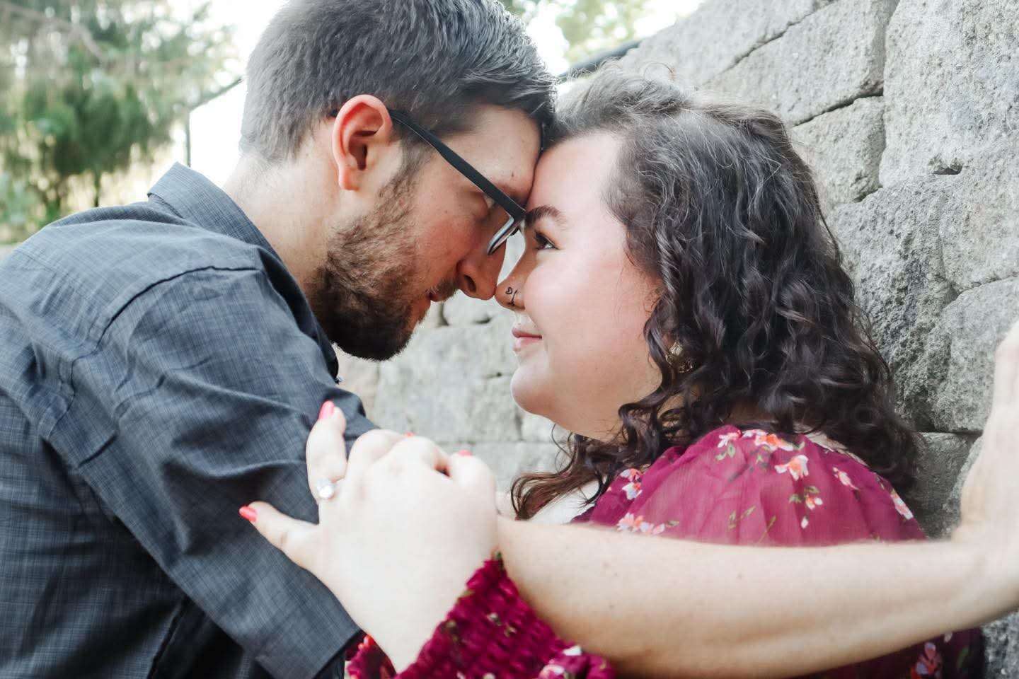 Couple leaning foreheads together against a stone wall, holding hands, woman in burgundy floral blouse