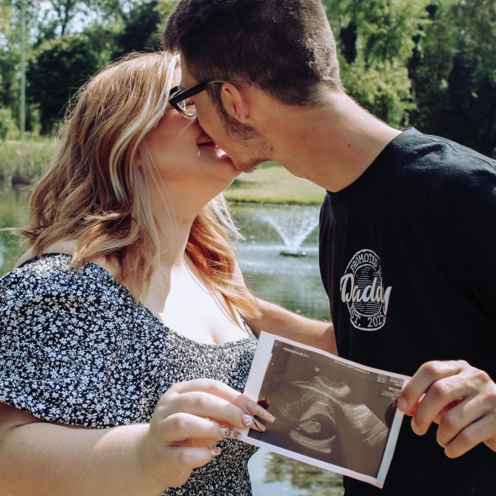 Couple kissing while holding their ultrasound photo in front of a park fountain, summer outdoor maternity session