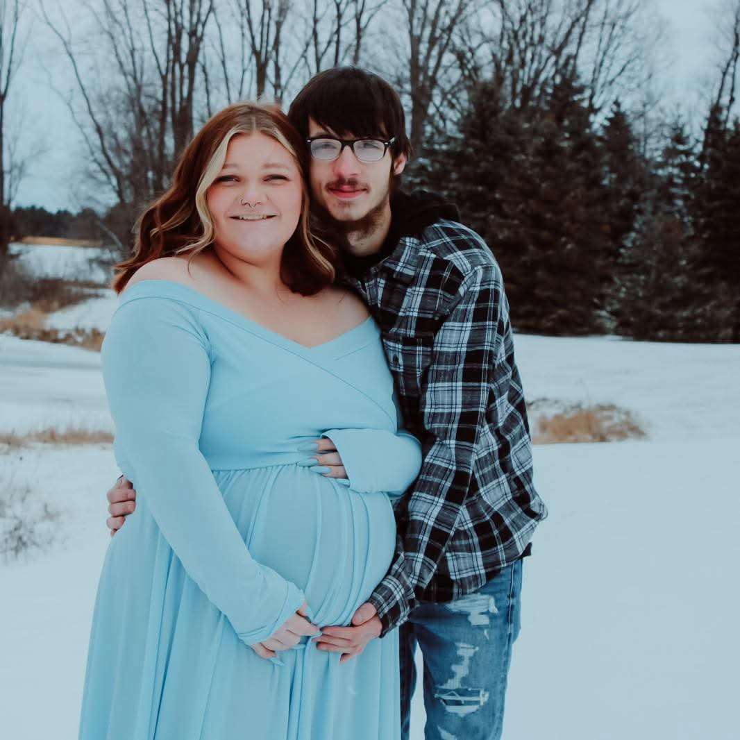 Couple embracing in a snowy outdoor setting, woman in a light blue off-shoulder maternity gown cradling her bump