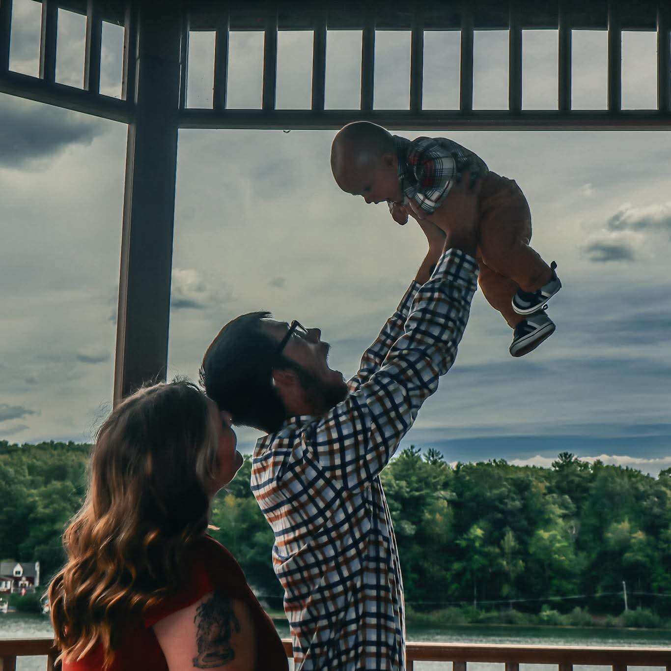 Father lifting baby high above his head on a covered lakeside deck, stormy sky and treeline behind them, mother watching with joy