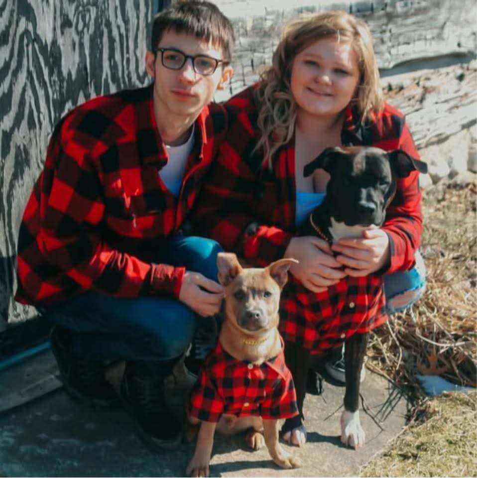 Couple in matching red buffalo plaid shirts crouching with their two dogs, tan puppy in a matching plaid shirt and a black dog