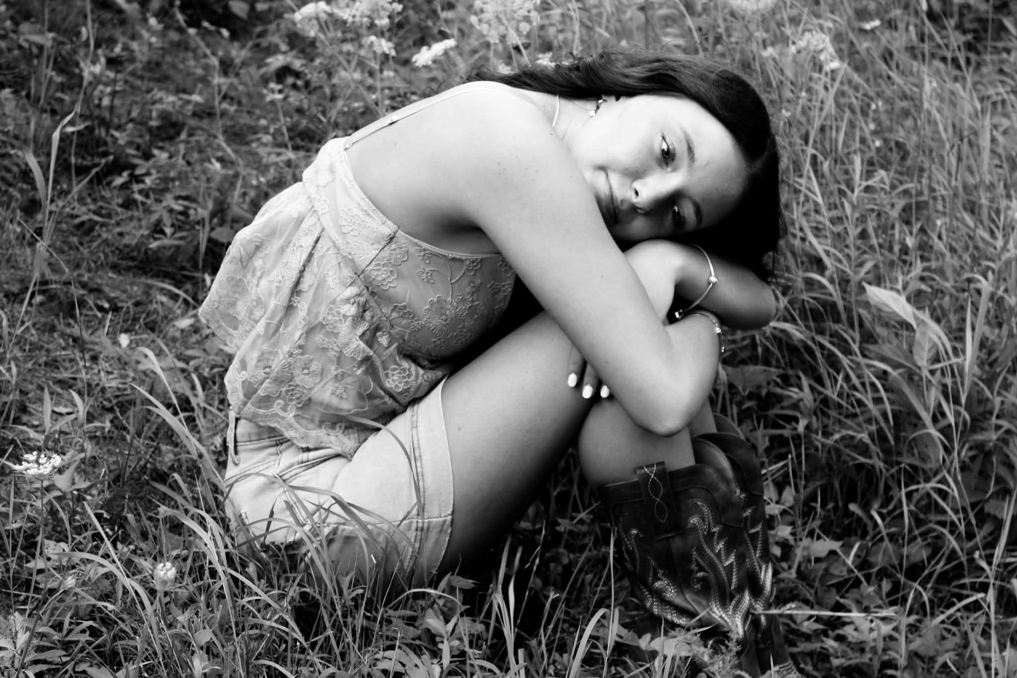 Black and white senior portrait of young woman in lace top and denim shorts with cowboy boots, sitting in a wildflower field