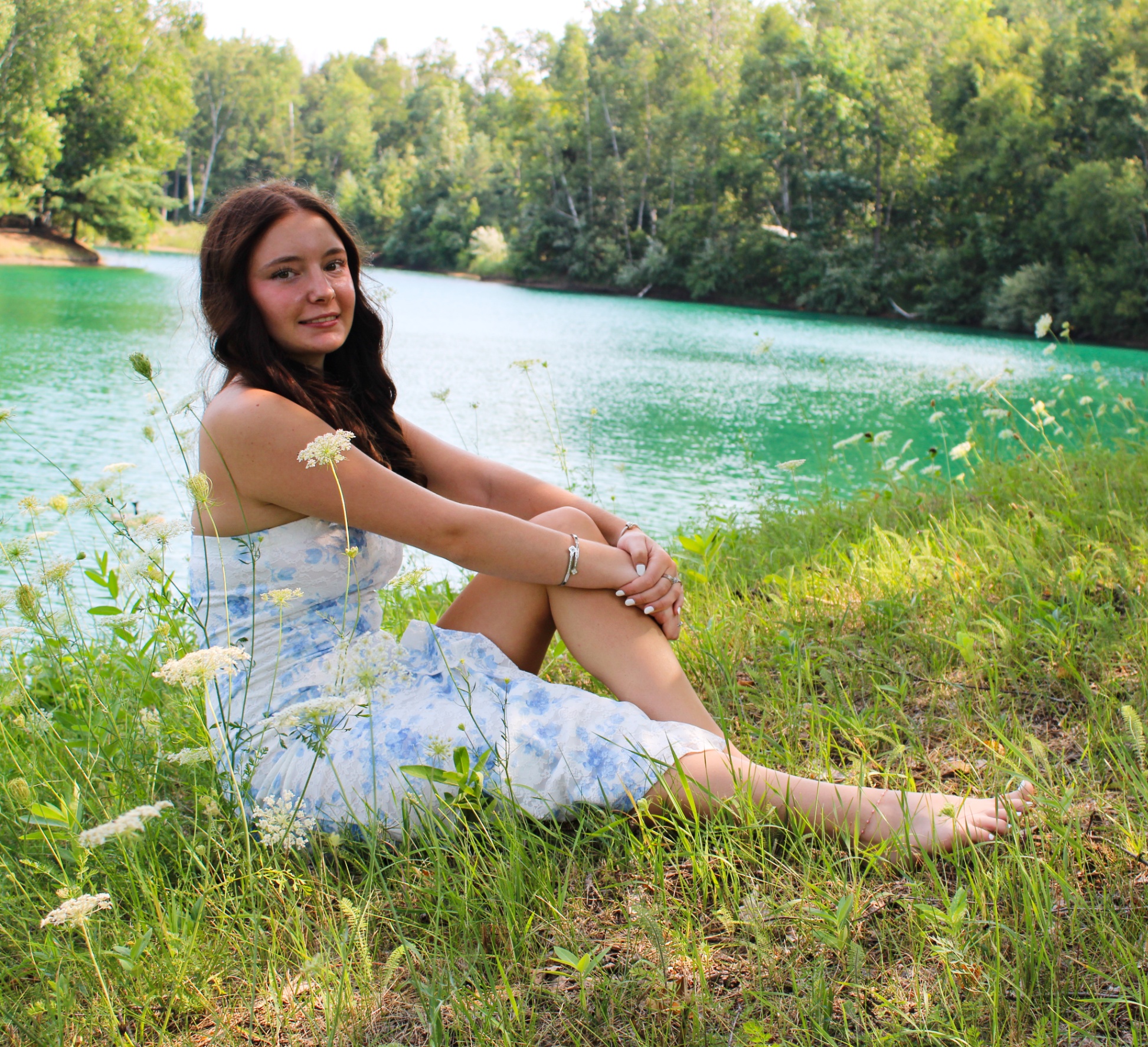 Young woman in a blue floral dress sitting relaxed on the grassy lakeside bank, lush green and turquoise water behind her