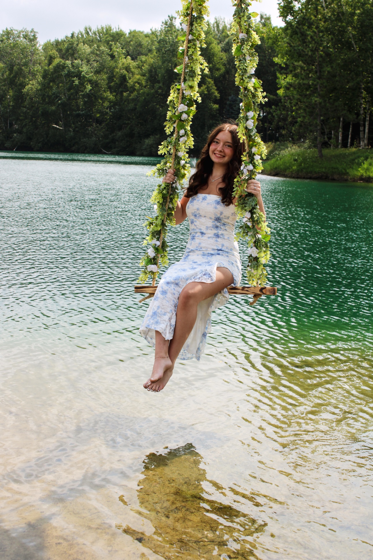 Young woman in a blue floral dress on a flower-vine swing over a turquoise lake, smiling at the camera, full body shot