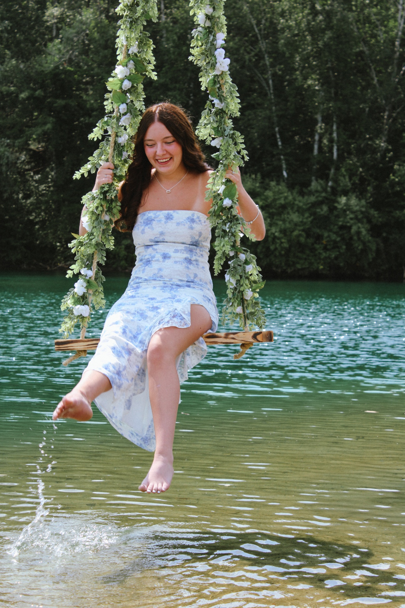 Young woman on a flower-vine swing over the lake, laughing and splashing water with her foot, candid and joyful