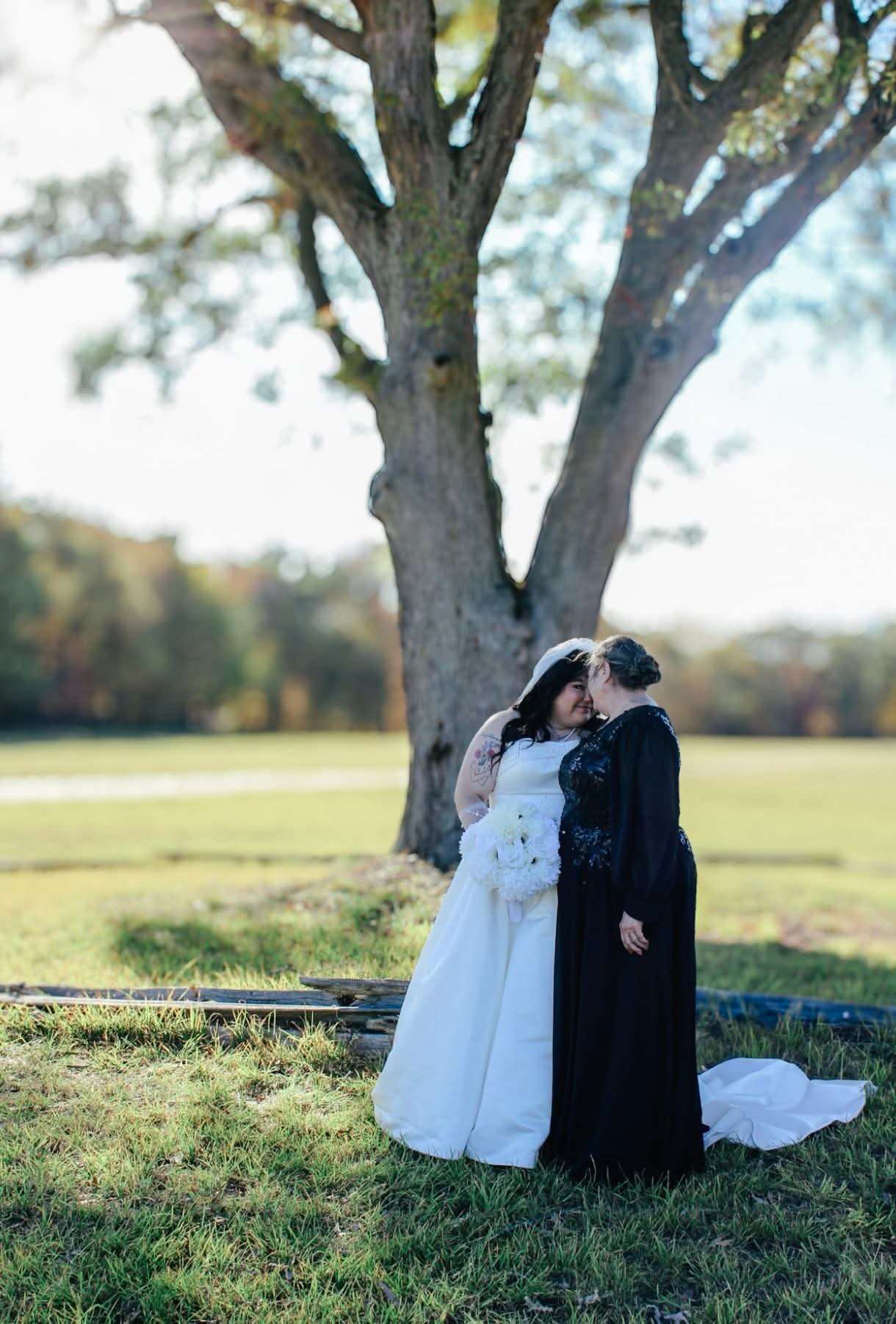 Two brides sharing an intimate moment under a large tree during a fall outdoor ceremony