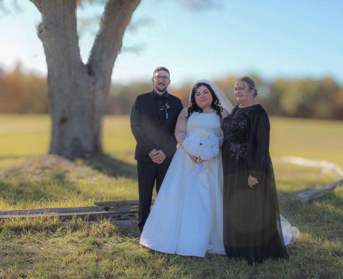Bride in white gown with groom in black tux and family member in black gown, golden hour light under a large tree
