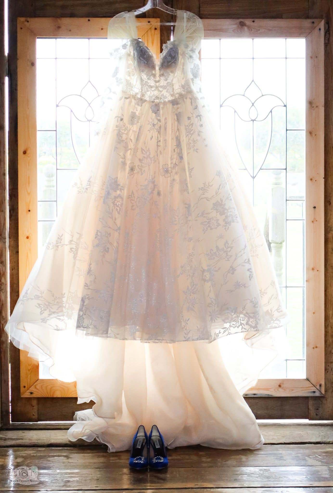 Wedding gown with blue floral embroidery hanging in a sunlit barn window, cobalt blue heels below